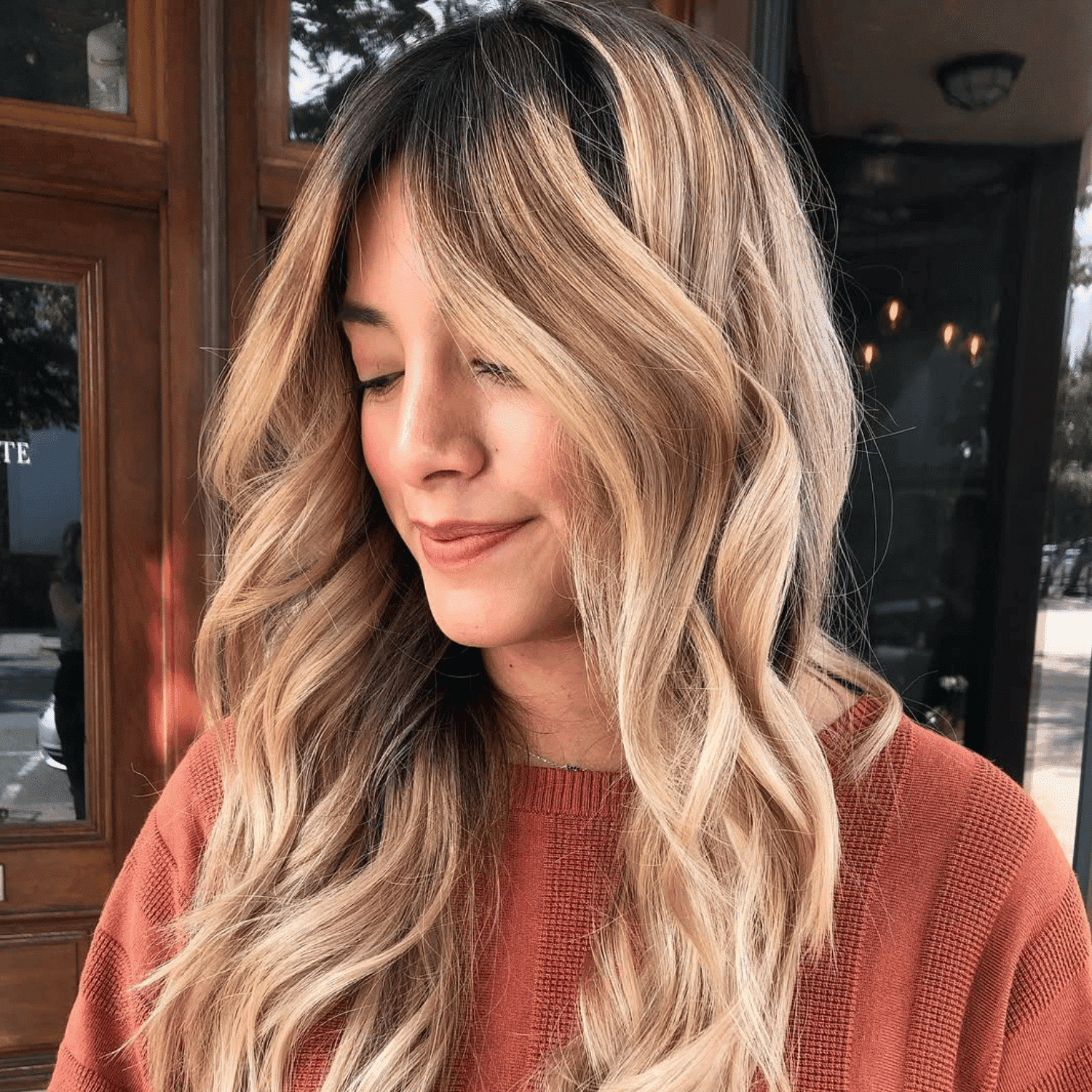 Woman with long, wavy blonde hair and a red sweater, smiling with eyes closed in satisfaction.